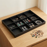 A black rectangular jewelry organizer tray with multiple compartments, shown holding assorted rings, earrings, bracelets, and necklaces on a light wood surface. A few loose jewelry pieces sit beside the tray.