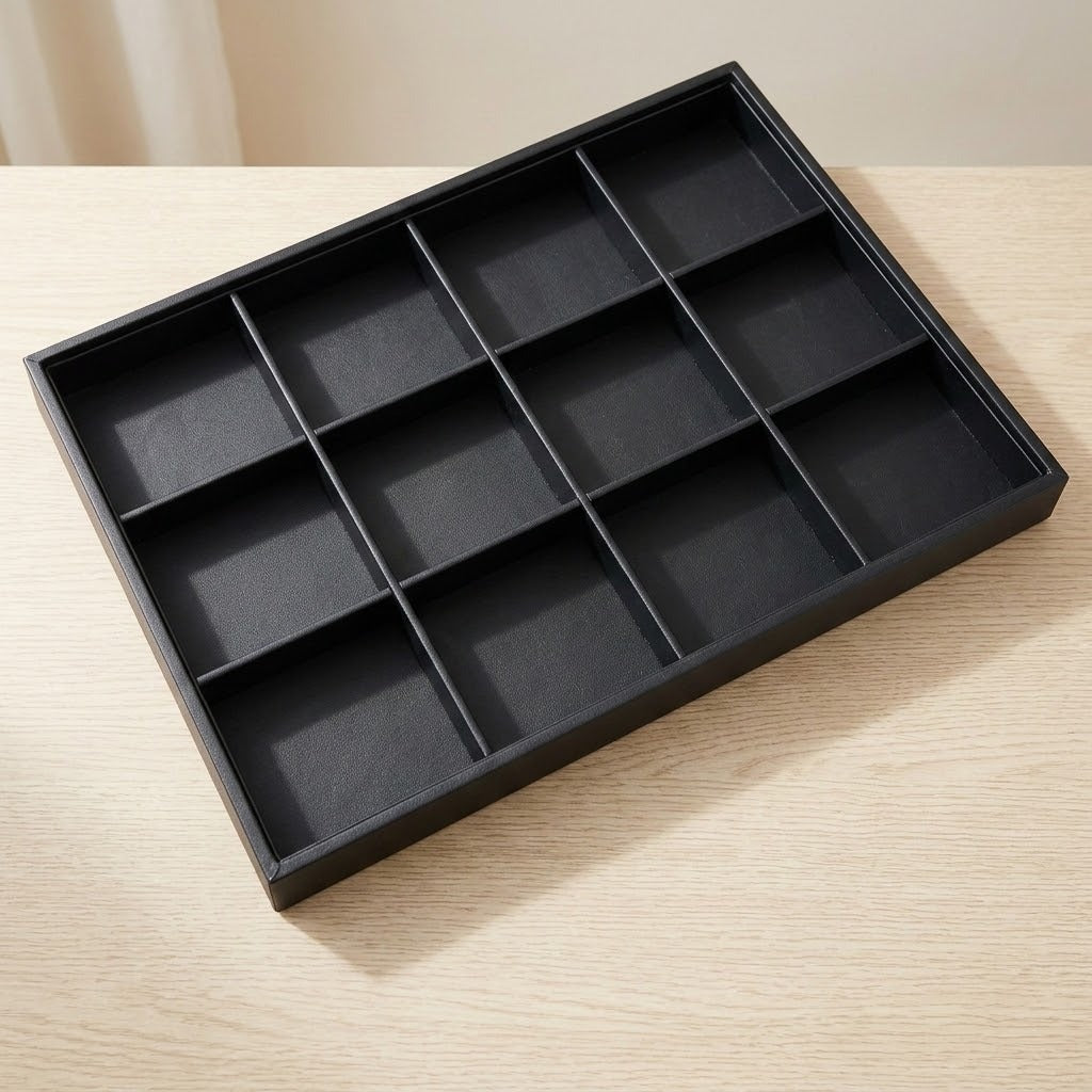 A black rectangular jewelry organizer tray with 12 individual compartments, shown on a light wood tabletop.
