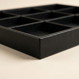 A black rectangular tray with nine square compartments, shown at an angle on a light beige background.