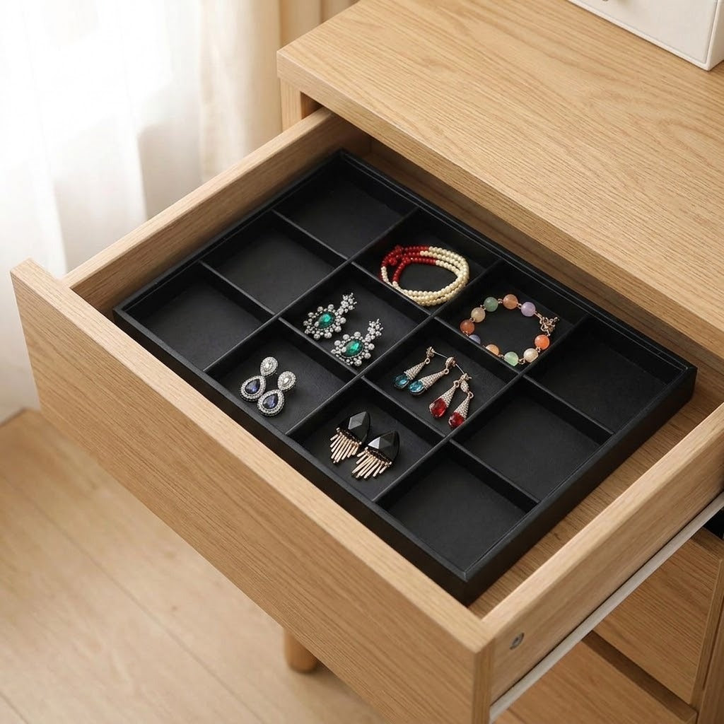A black drawer organizer tray with multiple compartments, shown inside an open light wood drawer containing assorted jewelry accessories.