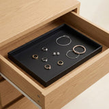 A black rectangular jewelry organizer tray placed inside an open light wood drawer, holding several rings, earrings, and bracelets.