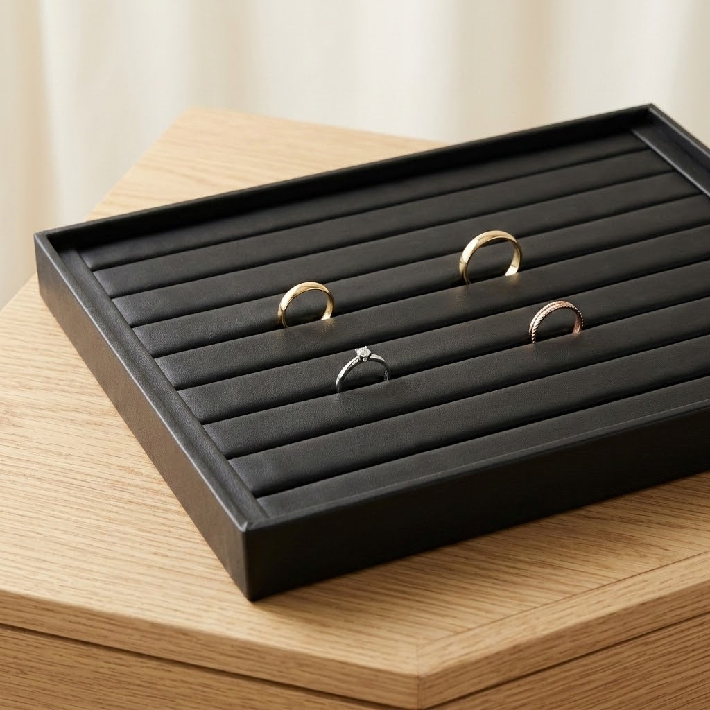 A black rectangular ring tray with padded horizontal slots, shown on a light wood surface. Three rings are displayed inside the tray, including two gold rings and one silver ring with a clear stone.
