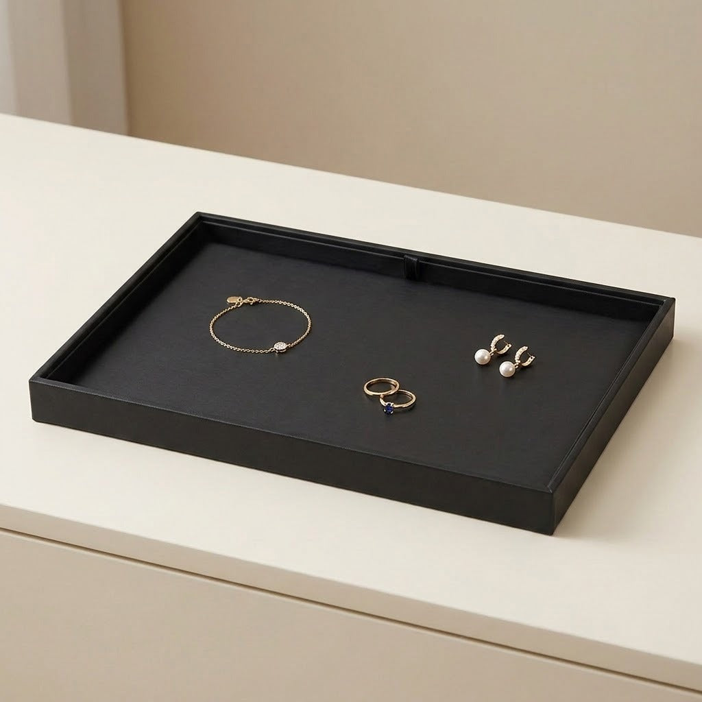 A black rectangular jewelry organizer tray with a textured black velvet-like interior, shown on a light cream surface. Inside the tray are several pieces of jewelry, including a gold bracelet, earrings, and rings.