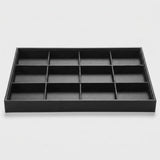 A black rectangular jewelry organizer tray with twelve individual compartments arranged in a 3x4 grid, shown on a light gray background.