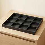 A black rectangular jewelry organizer tray with twelve compartments, shown resting on a light wood drawer or cabinet against a plain beige wall.