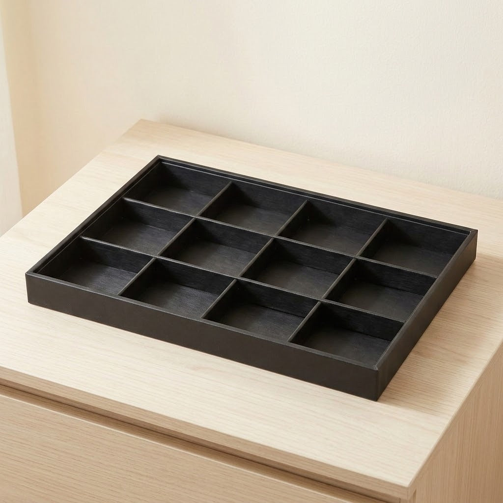 A black rectangular tray with nine divided compartments, shown resting on a light wood surface against a beige wall.