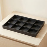 A black rectangular tray with nine divided compartments, shown resting on a light wood surface against a beige wall.