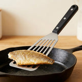 A silver slotted spatula with a black riveted handle lifts a browned fish fillet over a black skillet on a wooden counter.