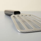 A stainless steel slotted spatula with a rectangular handle, shown in a close-up shot on a neutral gray surface with a warm beige background.