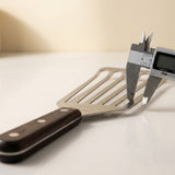 Digital calipers grip the silver edge of a slotted spatula with a black riveted handle on a white surface.