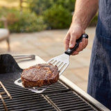 A person uses a silver slotted spatula with a black riveted handle to lift a grilled steak over an outdoor barbecue.