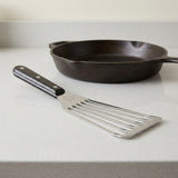A silver slotted spatula with a black riveted handle rests on a white countertop in front of a black skillet.