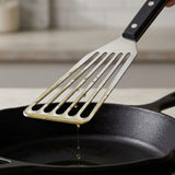 Oil drips from a silver slotted spatula with a black riveted handle held above a black skillet.