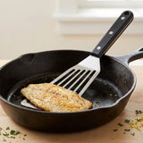 A black-handled silver slotted spatula sits beneath a browned fish fillet in a black skillet, with chopped herbs scattered on the wooden counter.
