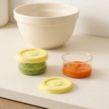 Two small clear baby food storage containers with yellow lids, one filled with green puree and the other with orange puree, shown on a white countertop with a large white bowl in the background.