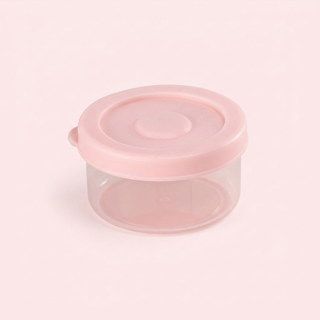 A small round plastic storage container with a translucent clear body and a light pink lid, shown on a pale pink background.