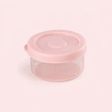 A small round plastic storage container with a translucent clear body and a light pink lid, shown on a pale pink background.