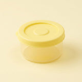A small round plastic storage container with a translucent yellow lid and a clear body, shown on a plain light beige background.