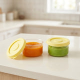 Two clear round containers hold orange and green purees on a white kitchen counter, with one yellow lid removed and one closed.