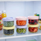 Six clear round containers with pink, yellow, and lavender lids hold fruit and vegetables, stacked in pairs on a refrigerator shelf.