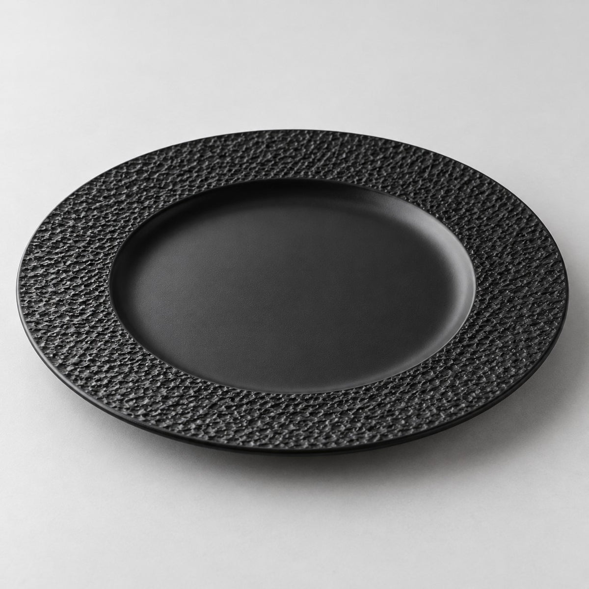 Black melamine dinner plate with a broad textured rim and smooth recessed center on a studio backdrop.