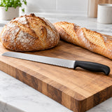 A stainless steel serrated bread knife lies beside a loaf and baguette on a kitchen cutting board.