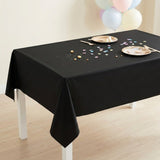 A black rectangular tablecloth draped over a table in a bright indoor setting, with plates, utensils, confetti, and pastel balloons visible in the background.