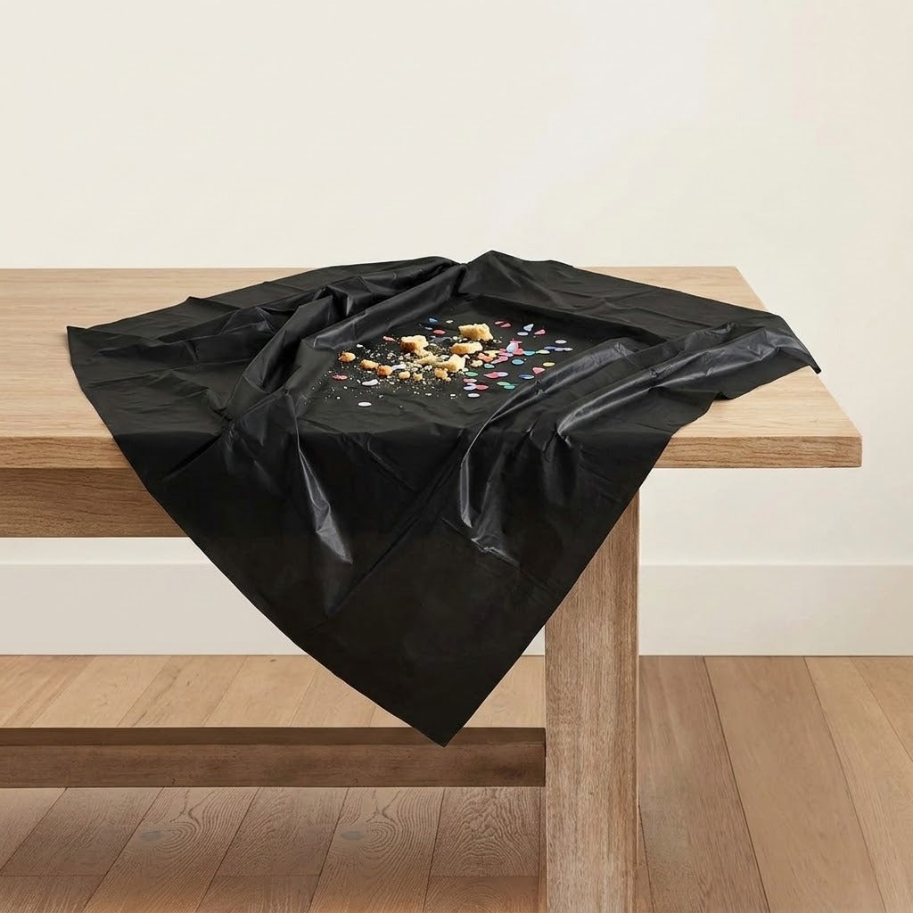 A black plastic tablecloth draped over a light wood table in a minimalist room setting with a cream wall and wooden floor.