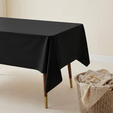 A black rectangular tablecloth draped over a table with wooden legs and gold-tone feet, set in a minimalist room next to a wicker basket holding a beige towel.