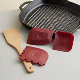Three red silicone scraping tools and a wooden spatula arranged on a light gray surface, with a black cast-iron grill pan in the background.