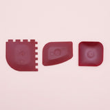 Three dark red silicone or rubber replacement parts with different shapes, laid out horizontally on a light pink background.