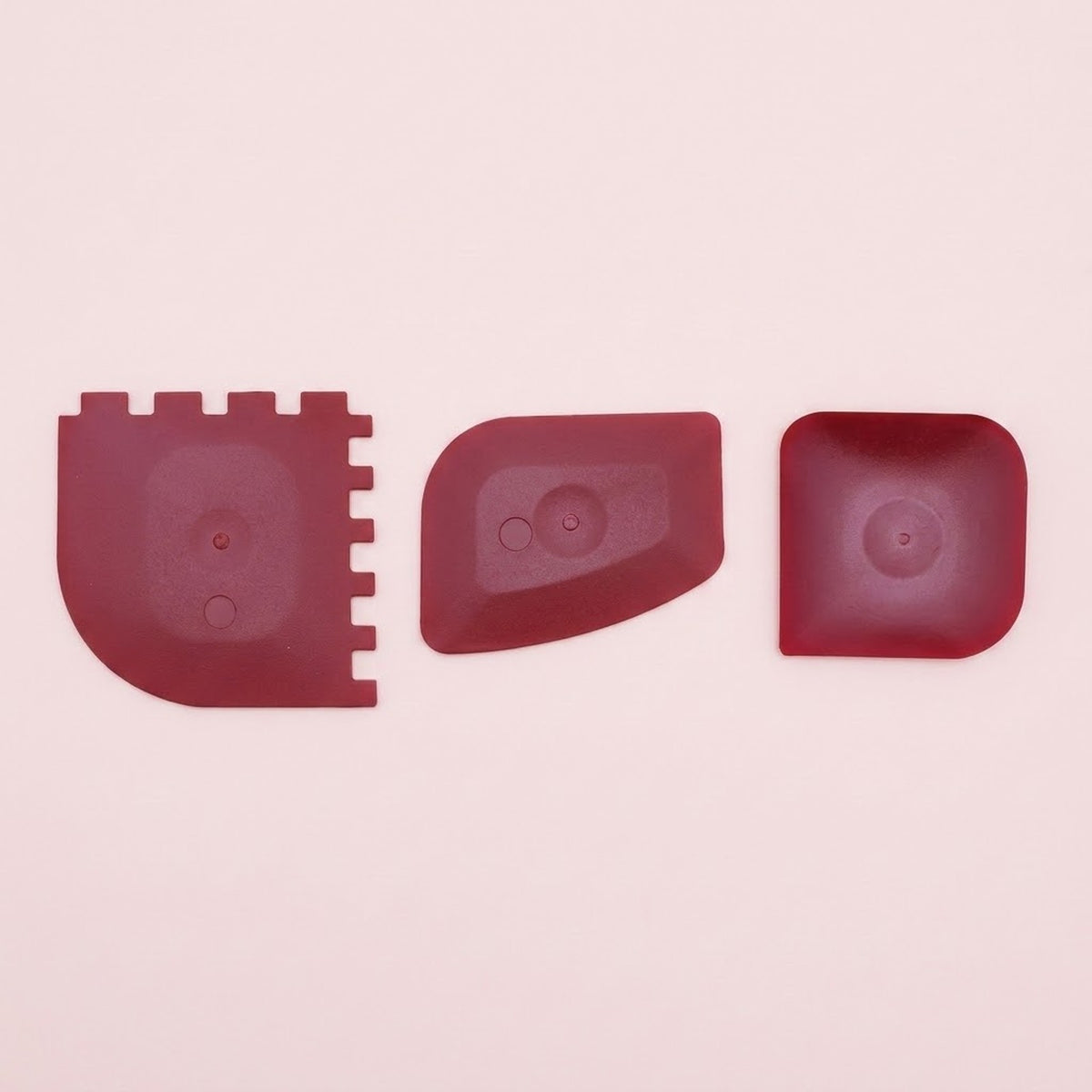 Three dark red silicone or rubber replacement parts with different shapes, laid out horizontally on a light pink background.