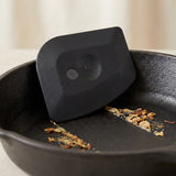 A black silicone pan scraper resting inside a black cast iron skillet, with some toasted food debris visible on the pan.