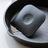 A black scraper with rounded corners rests inside a cast iron skillet beside loose food flakes on a pale countertop.