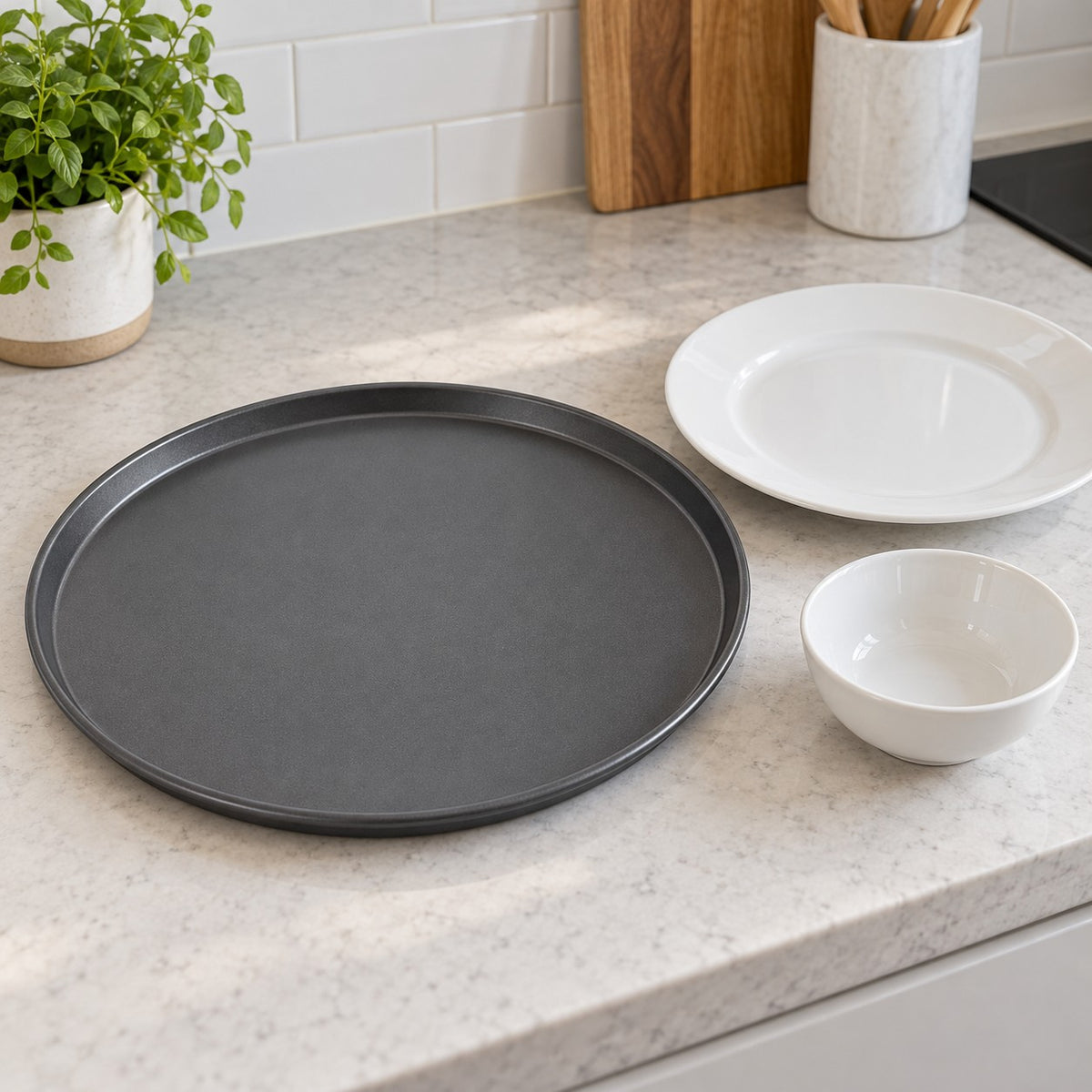 Round nonstick pizza pan beside a dinner plate and bowl on a kitchen counter for scale.
