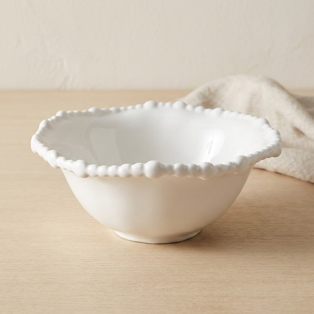 A white ceramic bowl with a scalloped, beaded rim, set on a light wood tabletop with a neutral beige background and a soft fabric napkin.