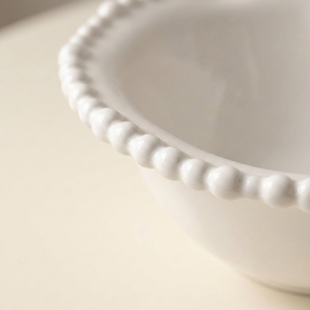 Close-up of a white ceramic bowl with a decorative beaded scalloped rim, set against a soft cream-colored background.