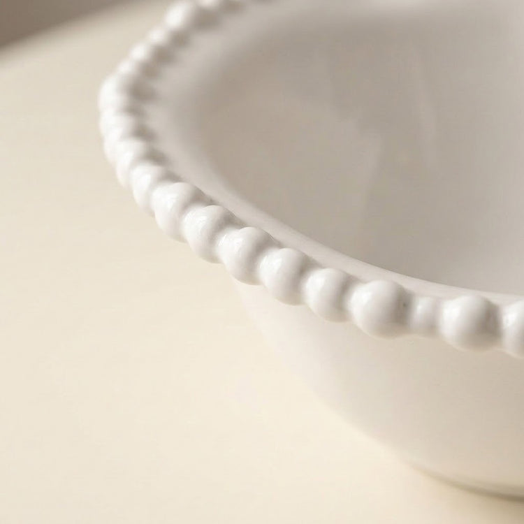 Close-up of a white ceramic bowl with a decorative beaded scalloped rim, set against a soft cream-colored background.