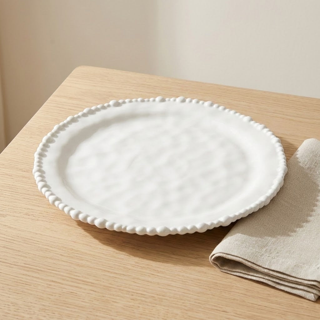 A round white ceramic plate with a textured center and a beaded rim, styled on a light wood table next to a folded beige cloth napkin.