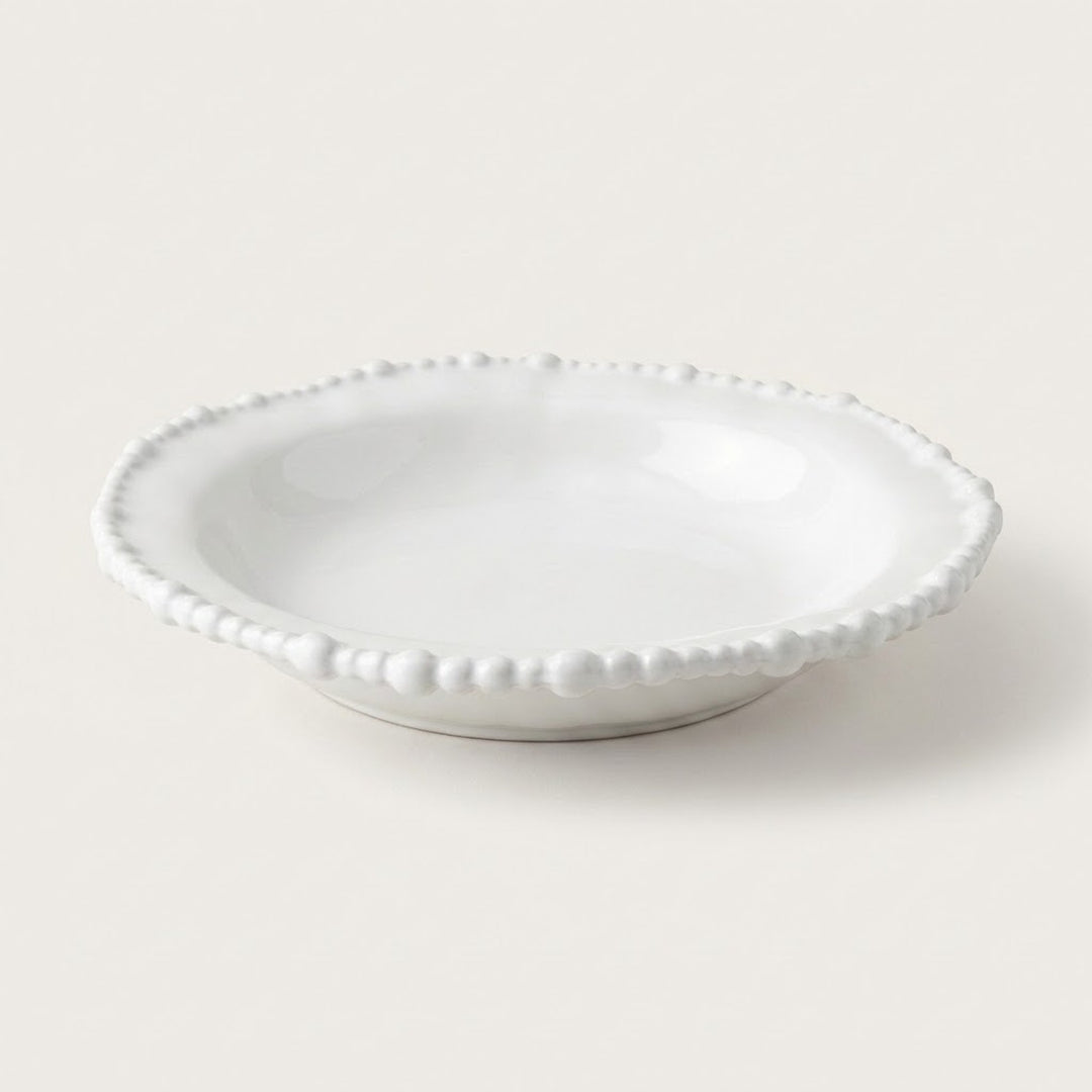 A small white ceramic dish with a scalloped beaded rim, photographed on a clean, solid, warm off-white background.