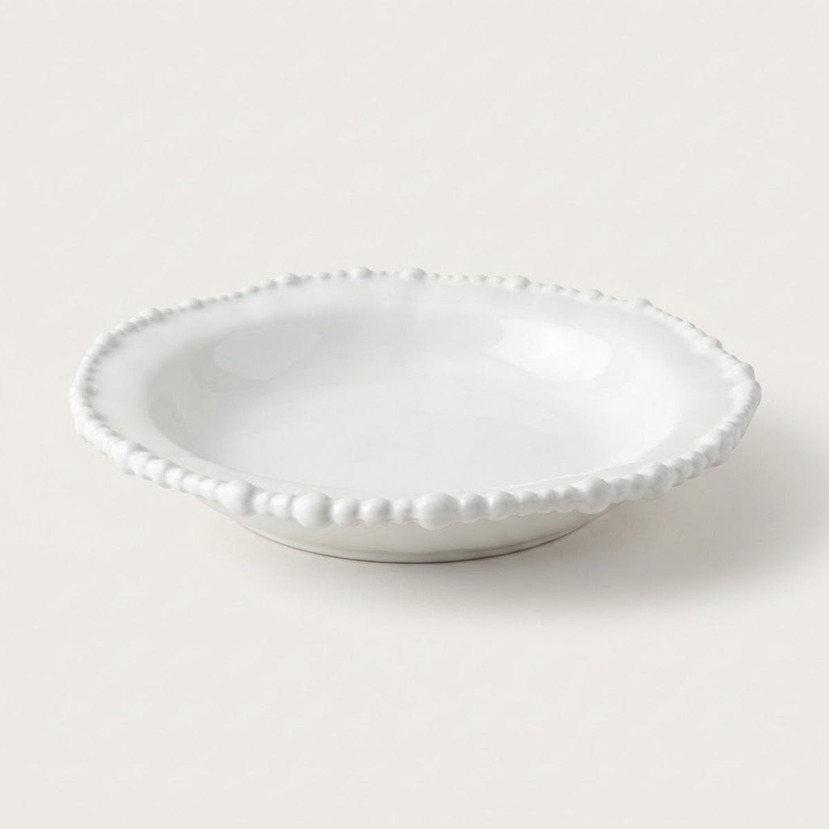 A small white ceramic dish with a scalloped beaded rim, photographed on a clean, solid, warm off-white background.