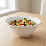 A white beaded-rim bowl holds lettuce, cucumber slices, cherry tomatoes, and shredded carrot on a wooden table near a window.