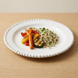 A white beaded-rim plate holds rice with herbs and roasted vegetables on a light wooden table.