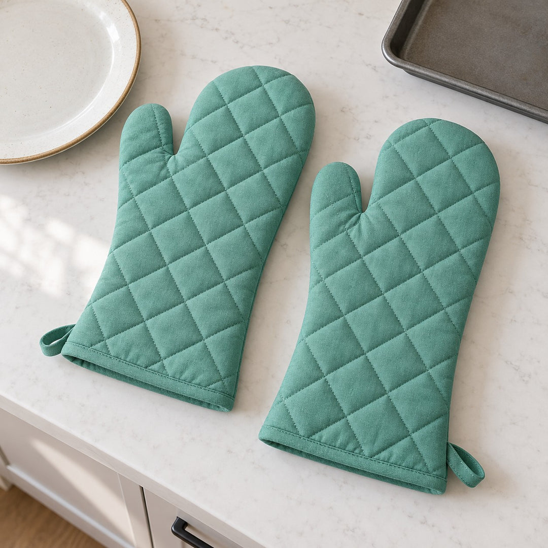 Turquoise oven mitt pair laid beside a dinner plate and baking tray to show their scale.