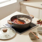 A two-tide hot pot set on a black induction cooktop, featuring a dark gray speckled pot with a wooden handle, a glass lid with a wooden knob, and a steaming red and milky broth inside. The scene is set in a modern kitchen with bowls, chopsticks, and ingredients nearby.
