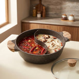 A split hot pot with one side containing spicy red broth and the other side containing creamy white broth, set on a light-colored table with a wooden kitchen background. A glass lid with a wooden handle is placed next to the pot.