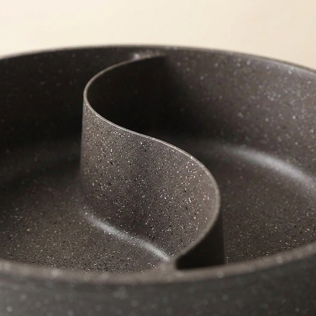 Close-up of a dark gray speckled non-stick divided pot or pan with a curved central divider, photographed against a light neutral background.