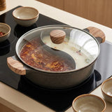A dark gray speckled hot pot with a clear glass lid and wooden handles sits on a black induction cooktop. The left side of the pot contains a red chili oil soup, while the right side has a light-colored broth.