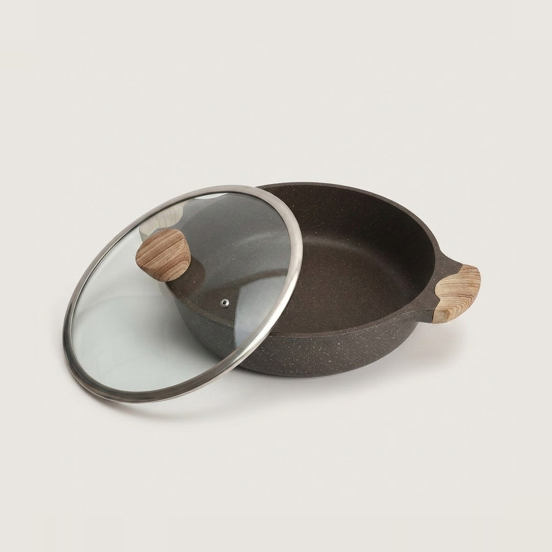 A dark speckled non-stick pan with a matching glass lid featuring a silver rim and light wood handles, shown on a clean off-white background.