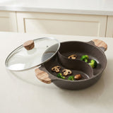 A dark gray speckled divided cooking pot with two wooden handles and a clear glass lid, shown on a light countertop with food pieces inside the compartments.